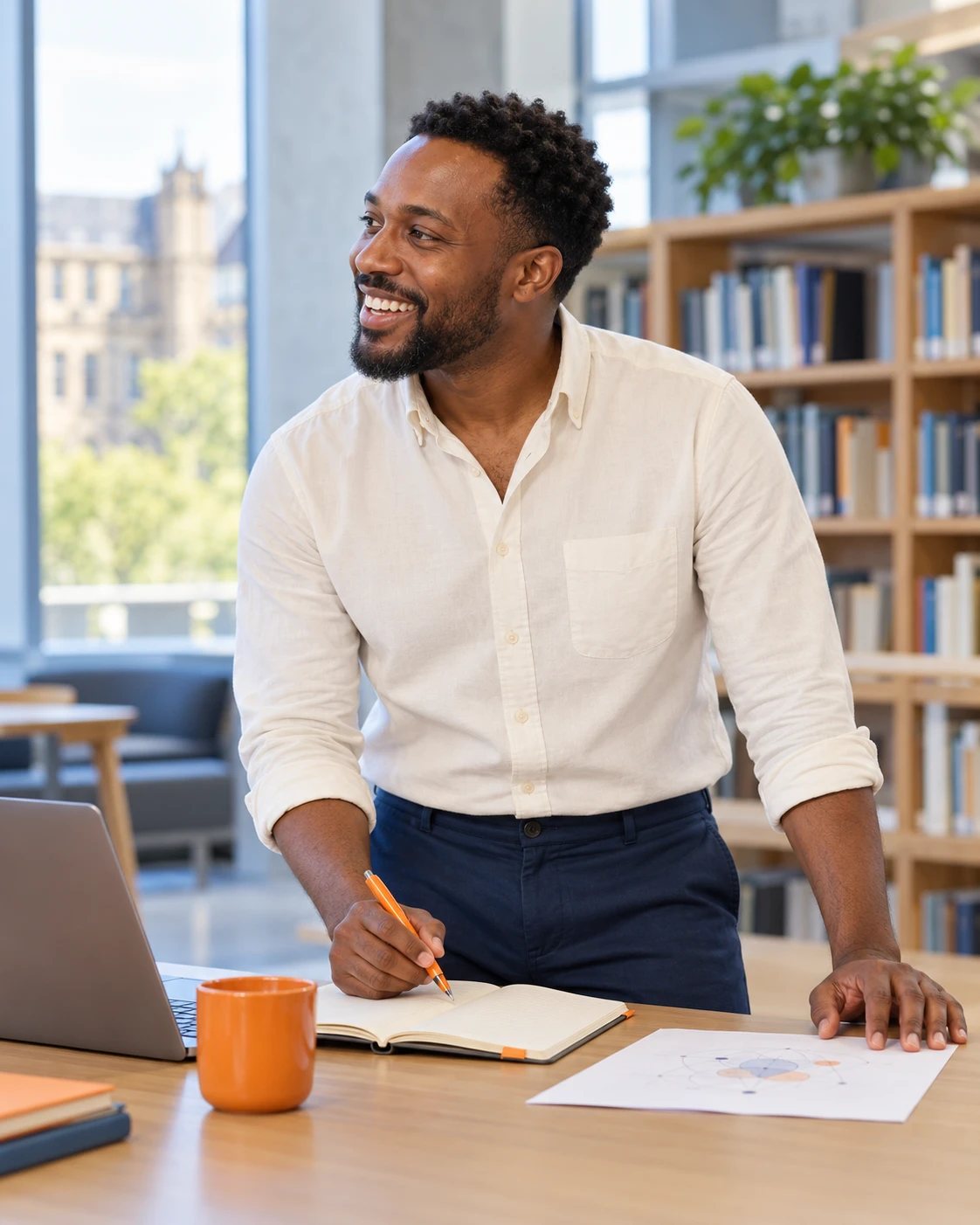 A smiling researcher standing at a desk in a bright university library, writing in a notebook with an orange pen